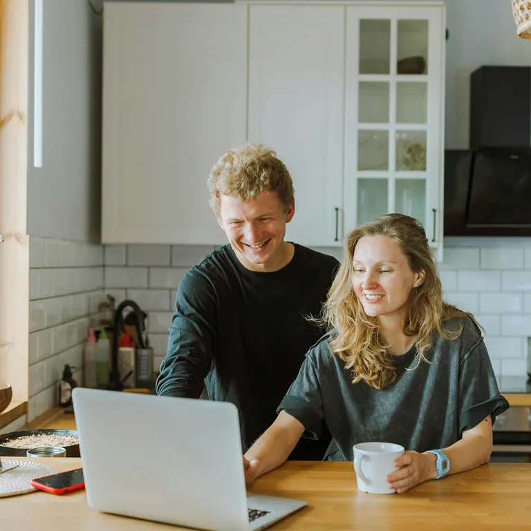 Ein junges Paar sitzt lächelnd in einer modernen Küche vor einem Laptop. Sie schauen gemeinsam auf den Bildschirm – ein Moment digitaler Alltagsorganisation oder gemeinsamer Planung.