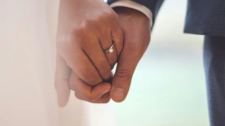 Detailaufnahme eines Paares, das sich an ihrem Hochzeitstag an den Händen hält. Im Fokus steht der Verlobungsring – Symbol für Liebe und Verbundenheit.